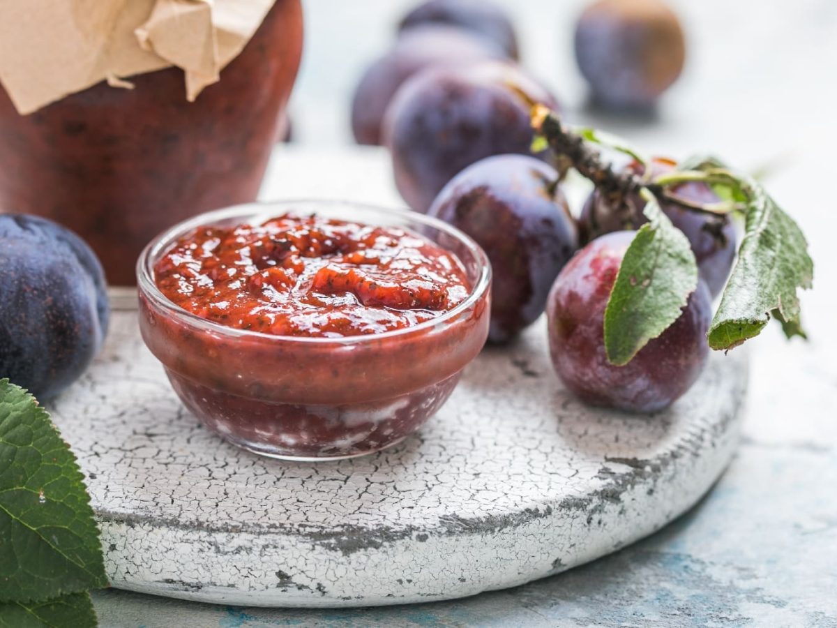 Eine kleine Glasschale mit der Pflauemnsauce auf einer grauen Steinplatte. Im Hintergrund auch frische Pflaumen und ein Glas mit Konfitüre.