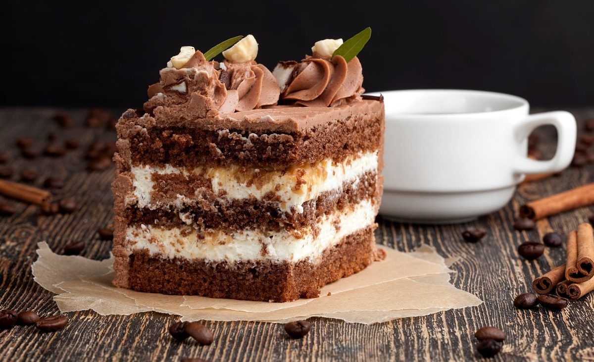 Frontalsicht: eine angeschnittene Torte aus Creme, Sahne und Schikolade steht auf einem dunklen Holztisch. Daneben liegen Zimtstangen und Kaffeebohnen. Und eine helle Tasse.
