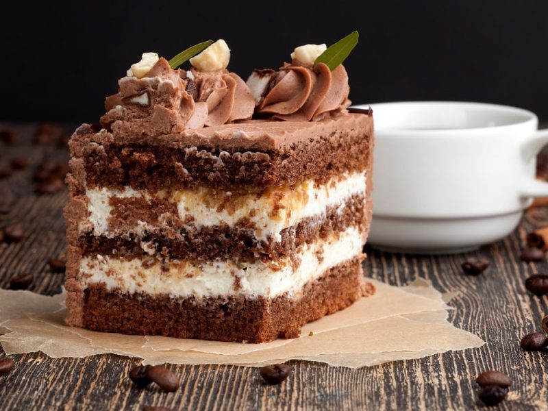 Frontalsicht: eine angeschnittene Torte aus Creme, Sahne und Schikolade steht auf einem dunklen Holztisch. Daneben liegen Zimtstangen und Kaffeebohnen. Und eine helle Tasse.