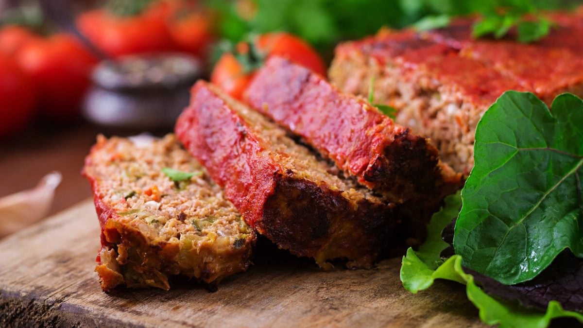 Ein angeschnittener veganer Hackbraten auf einem Brett, daneben frische Tomaten und Salatblätter.