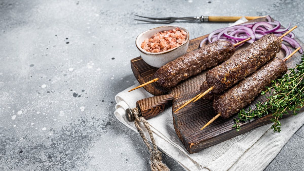 Drei Wild-Köfte-Spieße auf einem Holzbrett mit Thymian und Salz auf grauem Untergrund.