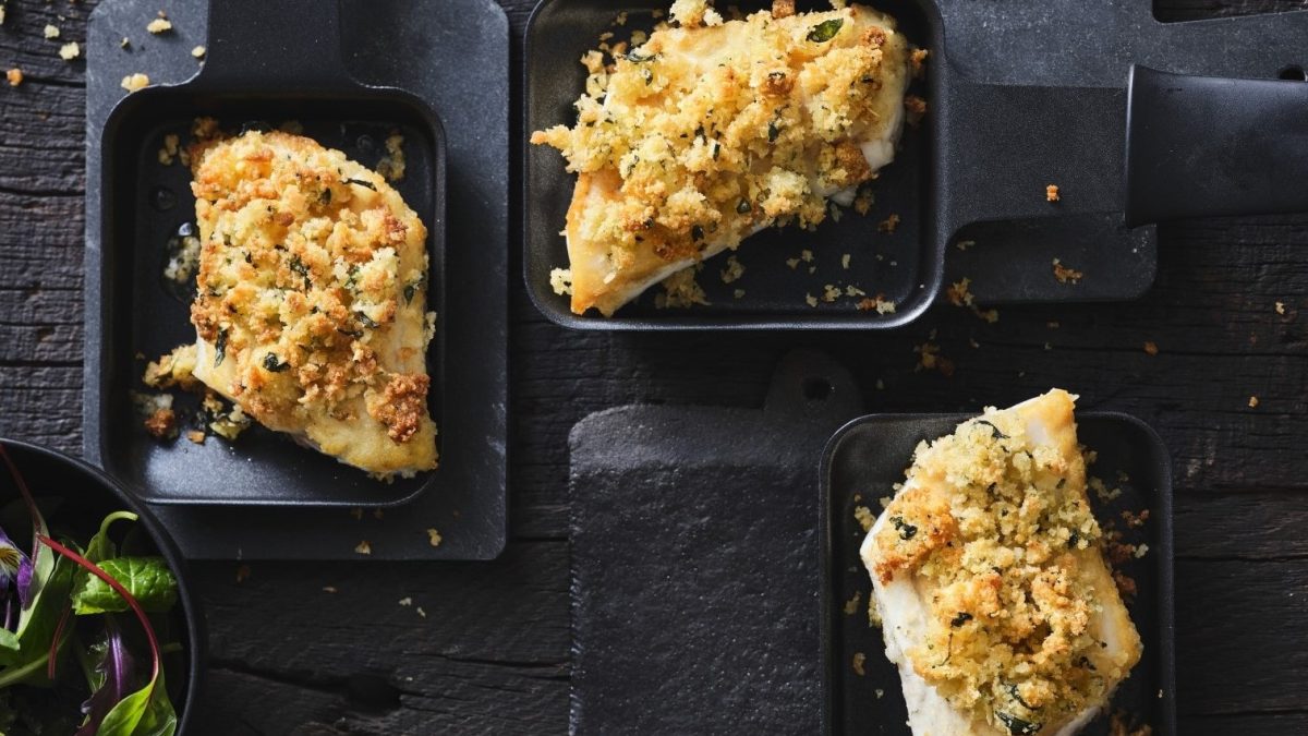 Wolfsbarsch mit Oregano-Parmesan-Kruste aus dem Raclette in drei Pfännchen auf schwarzem Untergrund. Draufsicht.