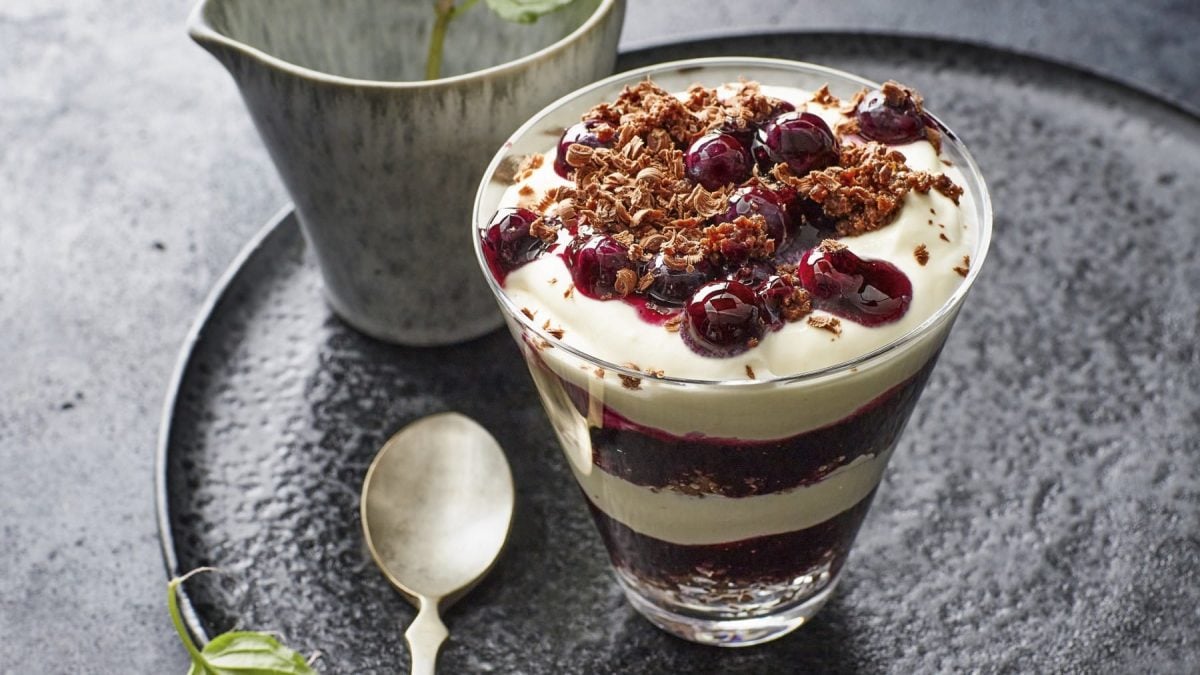 Stippmilch mit Blaubeerkompott und Schokoladenstreuseln, geschichtet mit dunkler Pumpernickelschokolade und serviert in einem durchsichtigen Dessertglas.