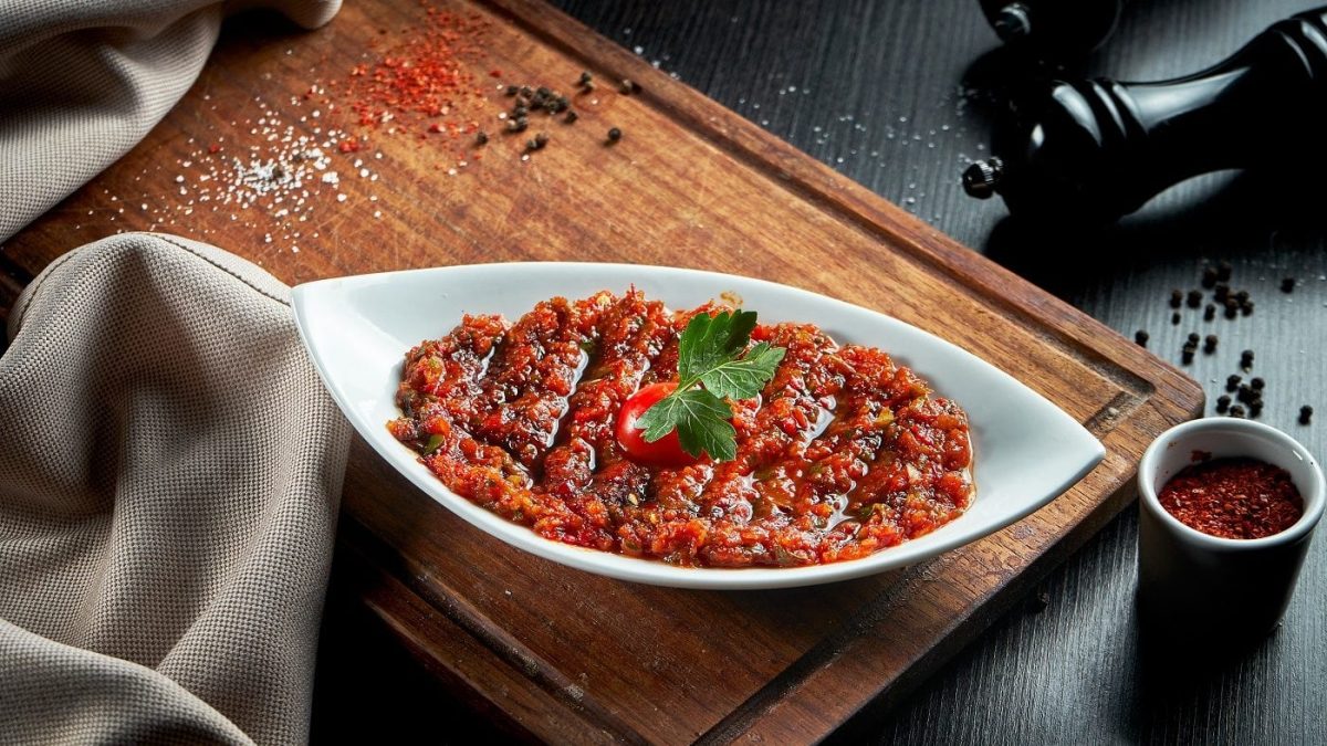 Draufsicht: Auf einem Holzbrett steht eine Porzellanschale mit der scharfen türkischen Gewürzpaste Acili Ezme. Drumherum liegen Kräuter und Gewürze.