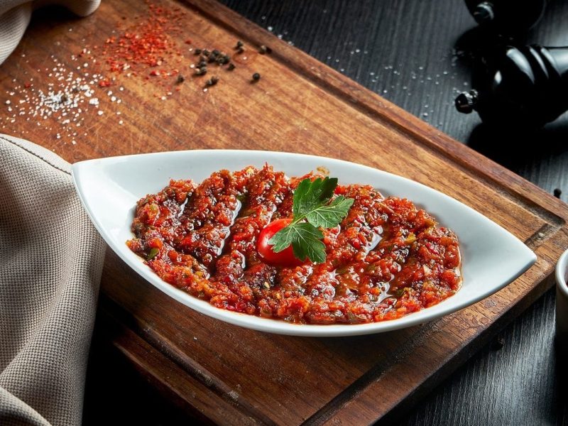 Draufsicht: Auf einem Holzbrett steht eine Porzellanschale mit der scharfen türkischen Gewürzpaste Acili Ezme. Drumherum liegen Kräuter und Gewürze.