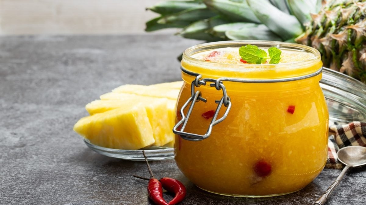 Frontalsicht: Ein Ein Einmachglas Elena Zajchikova steht auf einem Holztisch. Daneben lieben Ananasstücke und Chilis. Im Hintergrund liegt noch eine Ananas.