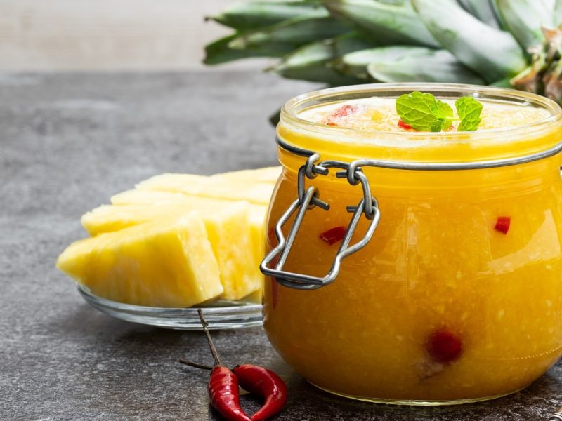 Frontalsicht: Ein Ein Einmachglas Elena Zajchikova steht auf einem Holztisch. Daneben lieben Ananasstücke und Chilis. Im Hintergrund liegt noch eine Ananas.