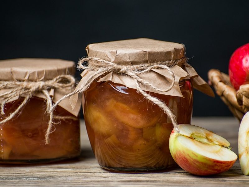 frontalsicht: zwei einweckgläse mit einem apfel-minz-relish stehen auf einem untergrund aus holz. der hintergrund ist dunkel. neben den gläsern, liegern halbierte und ganze äpfel.