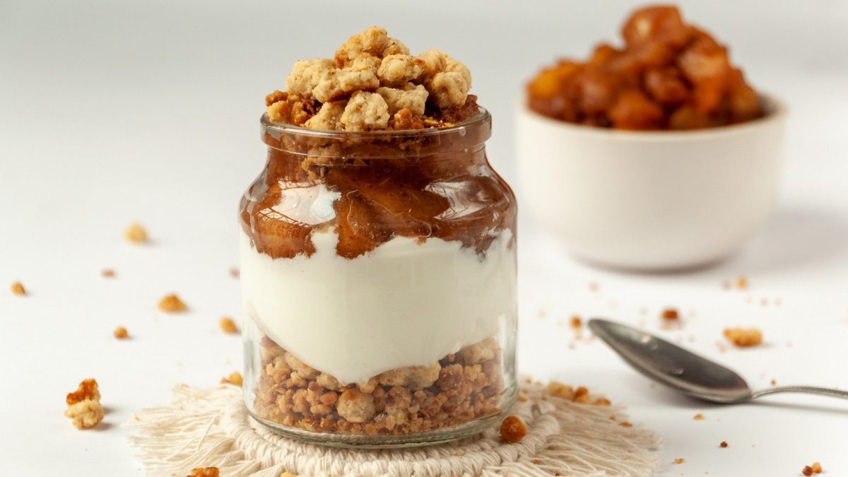 Ein Glas mit Apfel-Zimt-Tiramisu: unten Kekskrümel, darüber weiße Creme, dann Apfelkompott und noch mal Kekskrümel. Im Hintergrund eine Schale mit Apfelkompott.