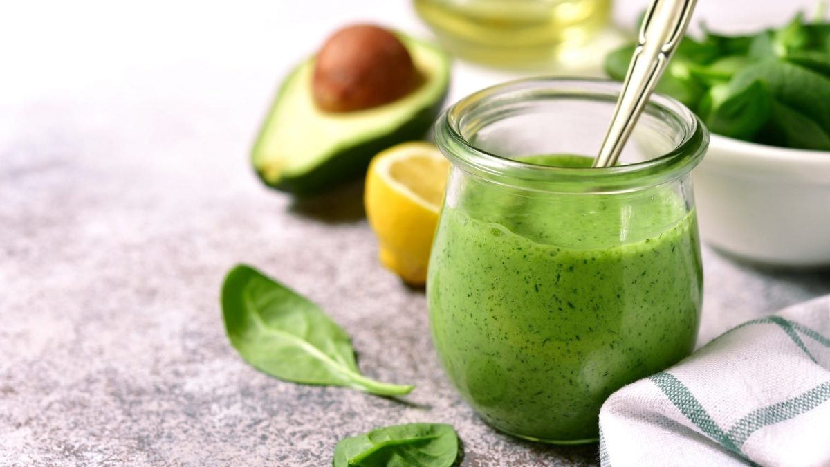 Ein Glas mit dem Avocado-Dressing auf einem grauen Tisch mit Zitrone, Babyspinat und Avocado.