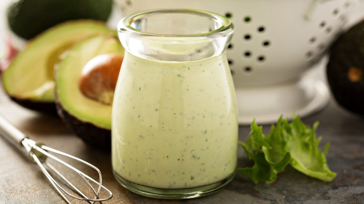 Eine kleine Glaskaraffe mit Avocado Ranch Dressing, dahinter unscharf eine halbierte Avocado, Salatblätter und ein Metallsieb.