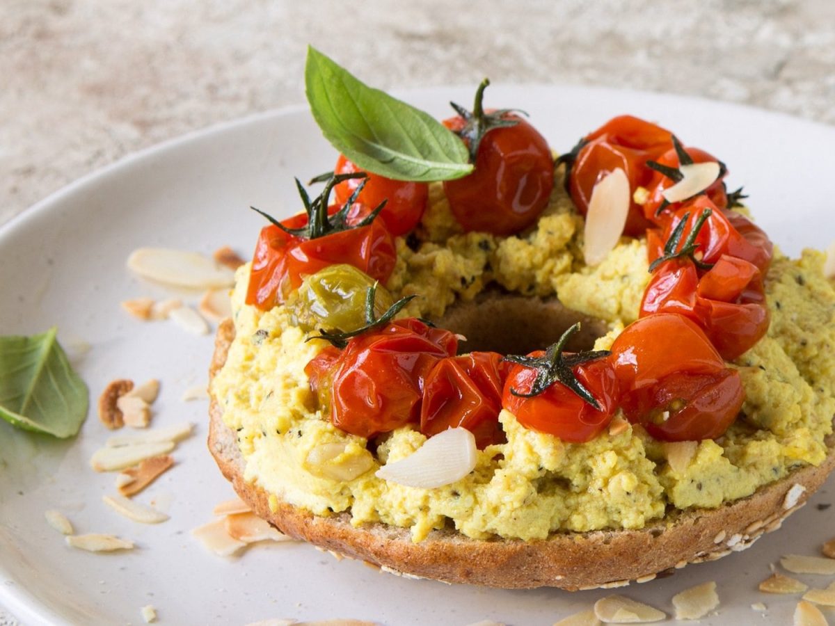 Ein Bagel-Sandwich mit Curry-Tofu, Basilikum, Mandelblättchen und Tomaten auf einem Teller.