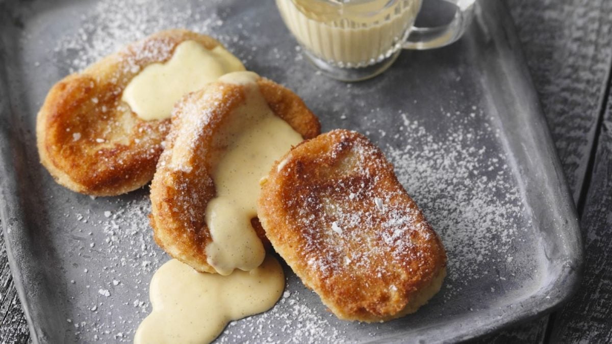 Draufsicht: Graues Tablett auf dem drei betrunkene Mönche mit Vanillesauce liegen. Bestäubt mit Puderzucker. Am oberen Bildrand steht eine Tasse mit zusätzlicher Vanillesauce.