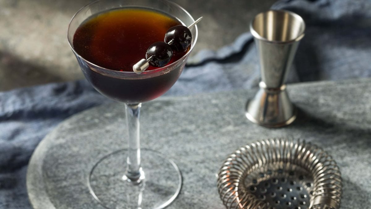 Black Manhattan Cocktail in Cocktailschale auf grauem Holztisch. Daneben Sieb und Barmaß. Draufsicht.