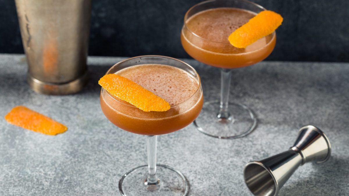 Zwei Boold and Sand Cocktail in Cocktailschale auf grauem Untergrund. Im Bild Barmaß, Shaker und eine Orangenzeste. Draufsicht.