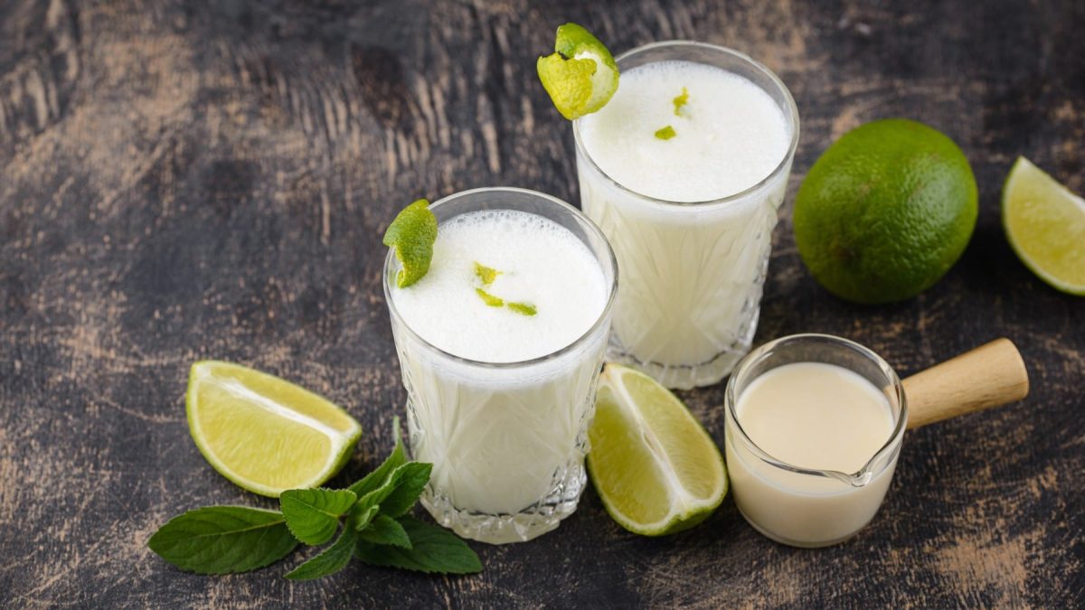 Brasilianische Limonade in zwei Gläsern, daneben Limettenviertel und Kondensmilch in einem kleinen Glas vor dunklem Holzhintergrund.
