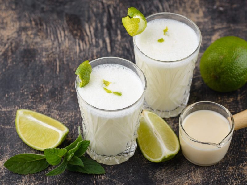 Brasilianische Limonade in zwei Gläsern, daneben Limettenviertel und Kondensmilch in einem kleinen Glas vor dunklem Holzhintergrund.