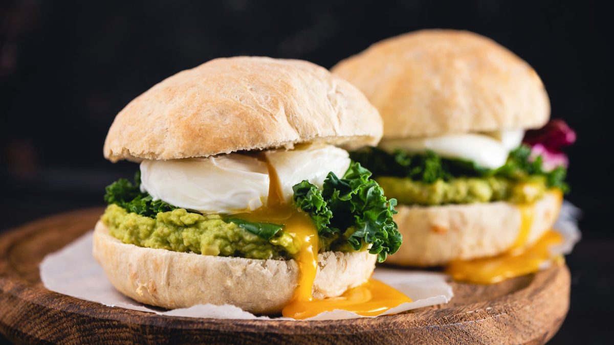 Breakfast-Burger mit pochiertem Ei, Schwarzkohl und Avocadosalsa im Brioche auf Krepp und dunklem Holz. Frontalansicht.