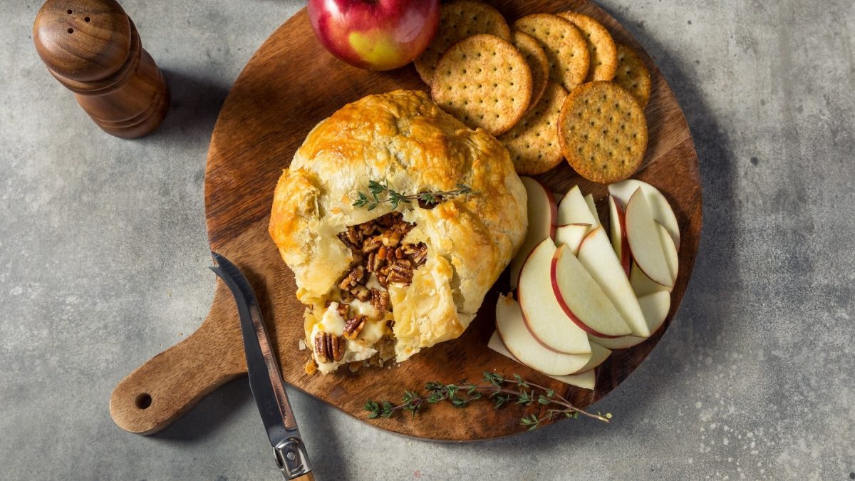 Brie-Pie mit Apfel und Pekannüssen gefüllt, angeschnitten serviert auf einem Holzbrett und angereicht mit Crackern und frischen Apfelscheiben.