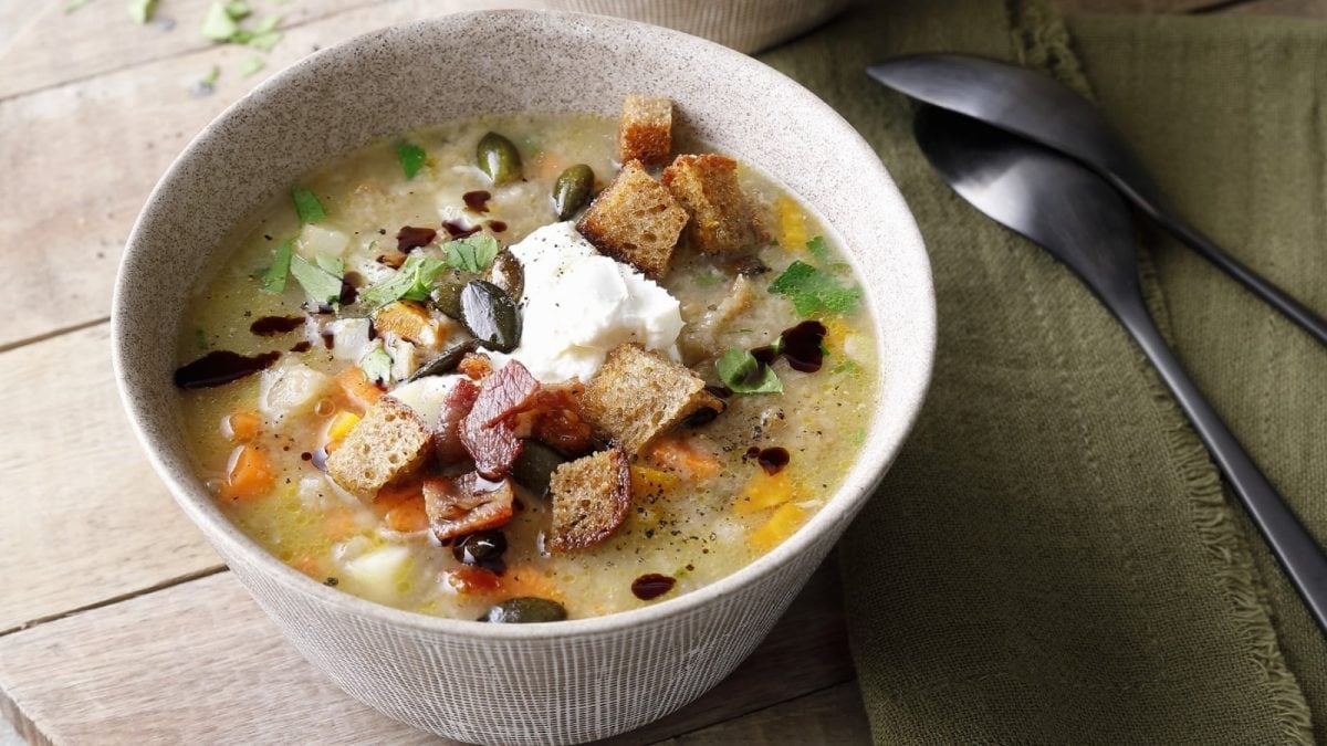 Draufsicht: Holzuntergrund. Auf der rechten Bildseite zwei silberne Löffel. Im Bildmittelpunkt eine Schüssel mit einer Suppe, getoppt mit Ziegenkäse, Kürbiskernen und Croûtons.