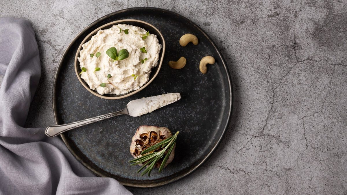 Eine Schüssel Cashew-Hummus auf einem Teller, darauf liegen außerdem Cashews und ein Messer sowie eine gebackene Knoblauchknolle. Draufsicht.