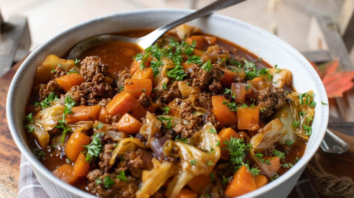 Eine weiße Schüssel mit dem Chinakohl-Eintopf auf einem dunklen Holztisch. Oben rechts ist ein silberner Löffel im Eintopf.