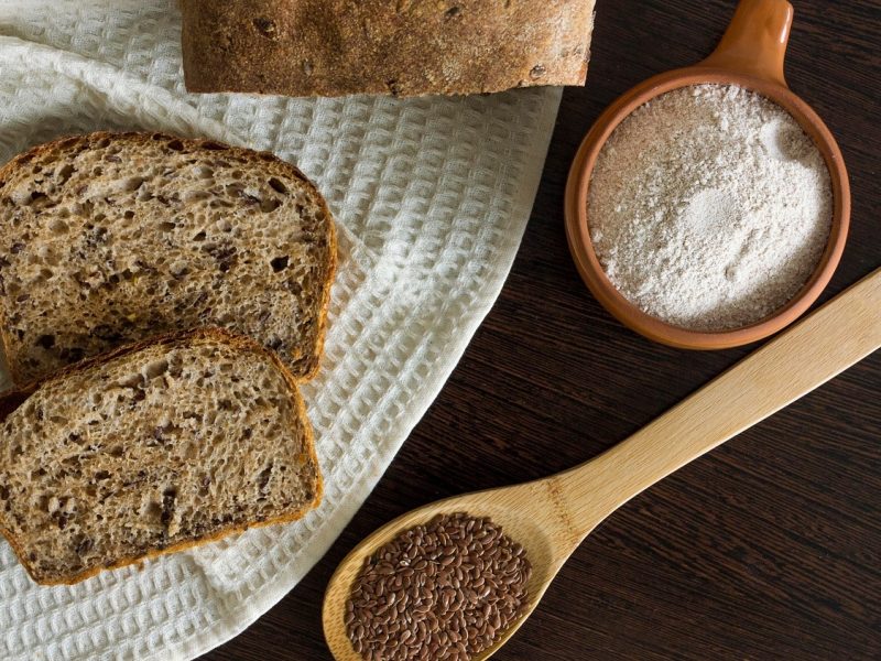 Draufsicht: Auf einem dunklen Untergrund ist ein kleiner Teil eines Leinsamenbrotes zu sehen. Daneben liegen zwei Scheiben Brot, ein Holzlöffel mit leinsamen und eine Schüssel mit Mehl.