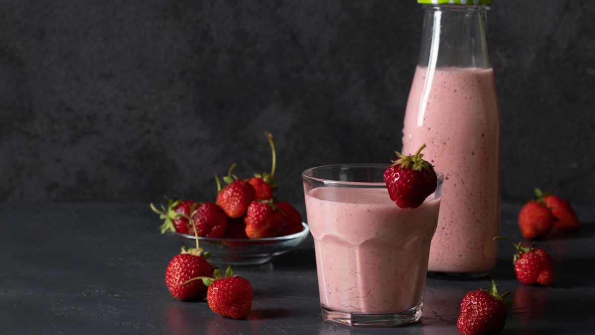 Erdbeer-Lassi im Glas, darum herum Erdbeeren und eine Flasche mit Lassi vor dunklem Hintergrund.