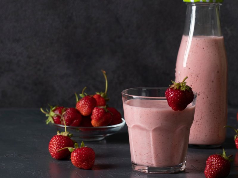Erdbeer-Lassi im Glas, darum herum Erdbeeren und eine Flasche mit Lassi vor dunklem Hintergrund.