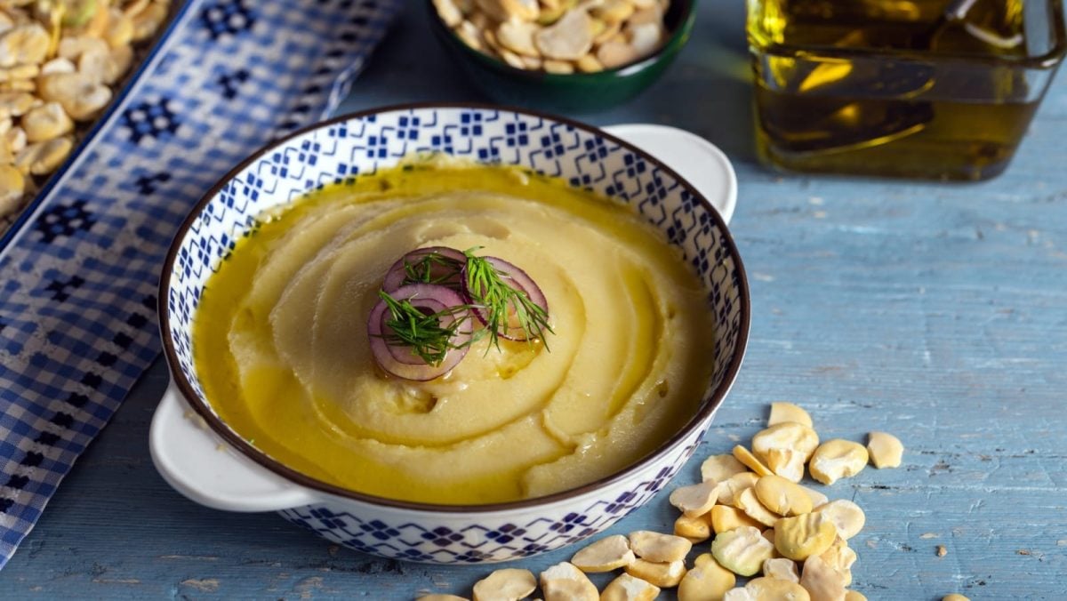 Blauer Untergrund mit einer Schüssel im Bildzentrum, welche mit einem griechisch-blau-weißem Muster bemalt ist. Darin ein cremiger Fava Aufstrich. Garniert mit frischer Petersilie. Um die Schüssel verteilt liegen lose Fava-Bohnen und frisches Olivenöl.
