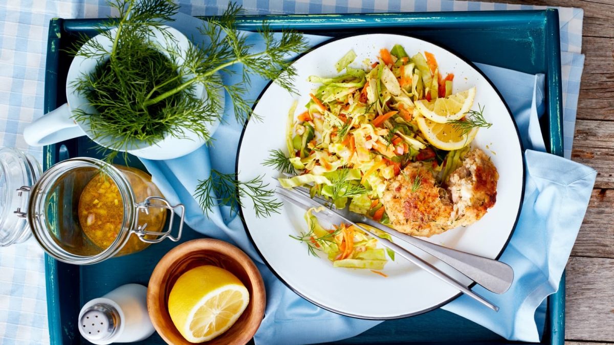 Draufsicht: Holztisch mit einem blauen Tablett auf dem auf der rechten Seite ein Teller mit Forellen-Frikadellen und Coleslaw steht. Auf der linken Seite ein Kännchen mit frischem Dill, ein Bügelglas mit Dressing und einer halben Zitrone, sowie einem Salzstreuer.