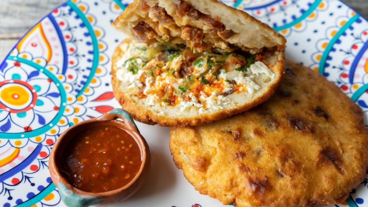 Zwei Gorditas de Chicharrón eines aufgeklappt mit einer Schale Salsa auf buntem Teller. Draufsicht.