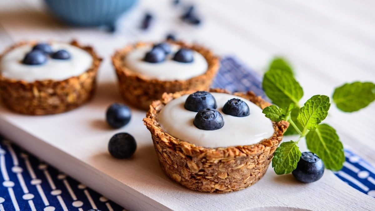 Frontalsicht: Auf einem hellen Untergrund liegt ein blaues, gemustertes Handtuch. Auf dem Handtuch liegt ein helles Holzbrettchen mit drei Granola-Cups mit Joghurt und heidelbeeren. Drumherum liegen frische Minzblätter.