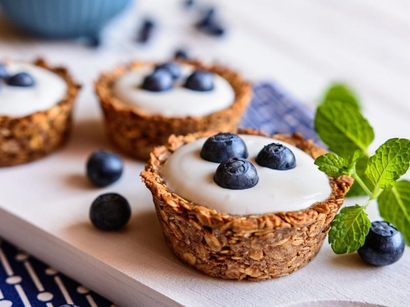 Frontalsicht: Auf einem hellen Untergrund liegt ein blaues, gemustertes Handtuch. Auf dem Handtuch liegt ein helles Holzbrettchen mit drei Granola-Cups mit Joghurt und heidelbeeren. Drumherum liegen frische Minzblätter.