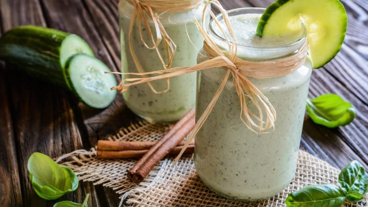 Zwei Gläser mit dem Gurken-Smoothie auf einem dunklen Holztisch. Daneben Gurke, Basilikum und Zimtstangen.