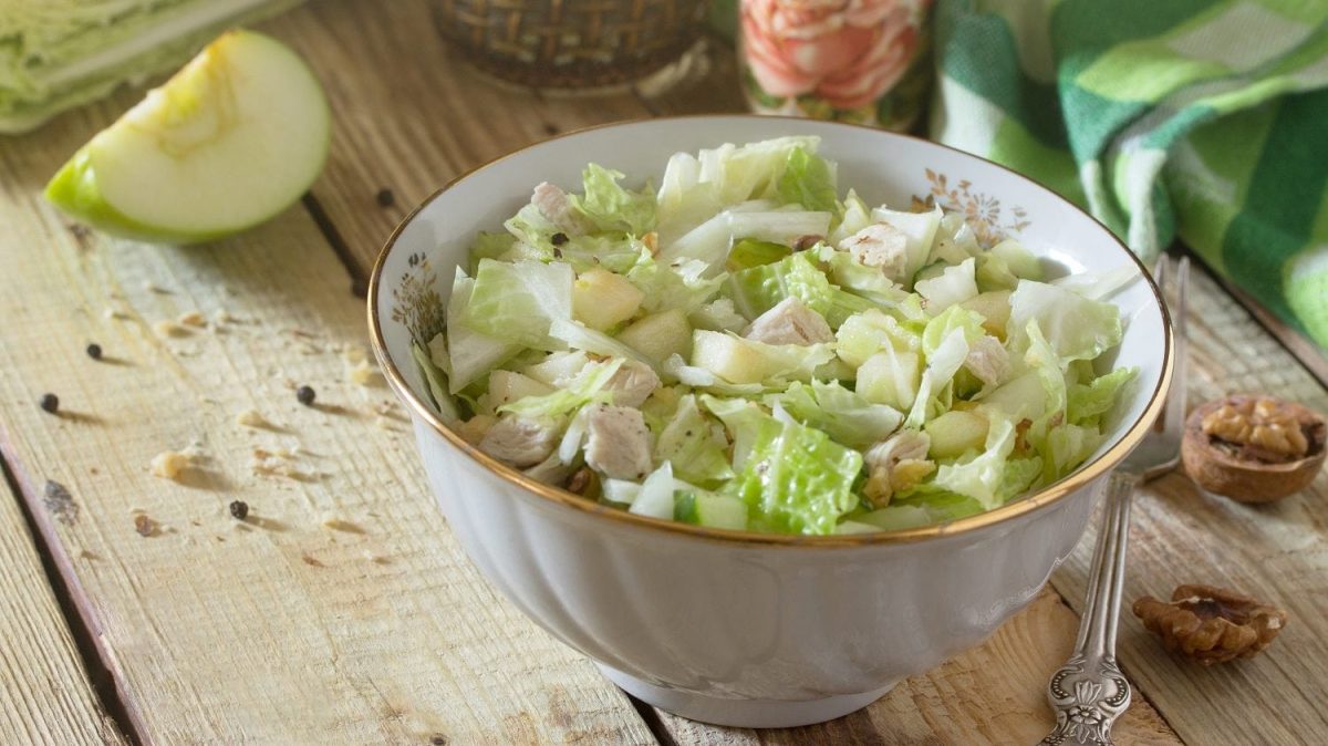 Schräge Draufsicht. Auf einem Untergrund aus Holz steht eine Porzellanschüssel mit einem Hühnchen-Apfel-Salat. Drum herum liegen Walnüsse, Gewürze sowie ein halber Apfel.