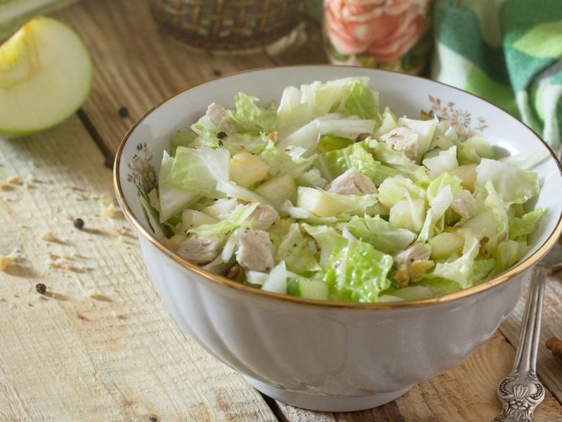 Schräge Draufsicht. Auf einem Untergrund aus Holz steht eine Porzellanschüssel mit einem Hühnchen-Apfel-Salat. Drum herum liegen Walnüsse, Gewürze sowie ein halber Apfel.