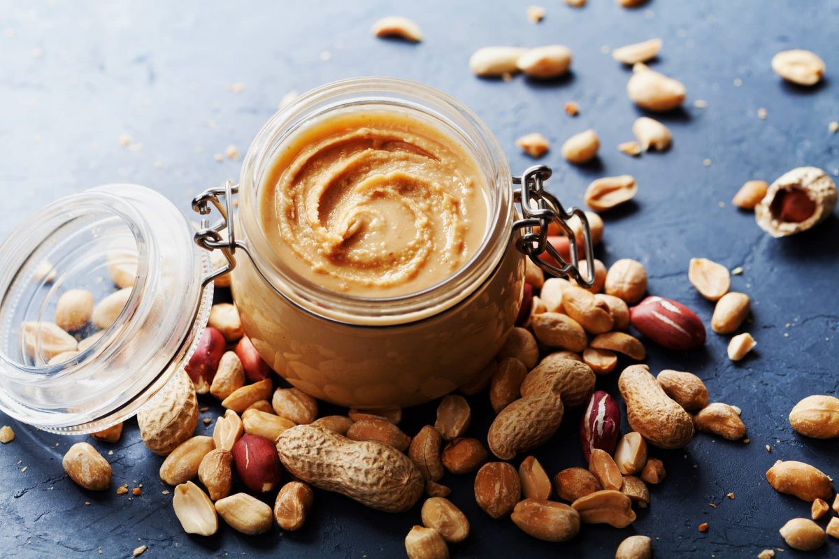 Ist Erdnussbutter vegan:: Ein Weckglas Erdnussbutter, drumherum ganze und geschälte Erdnüsse.