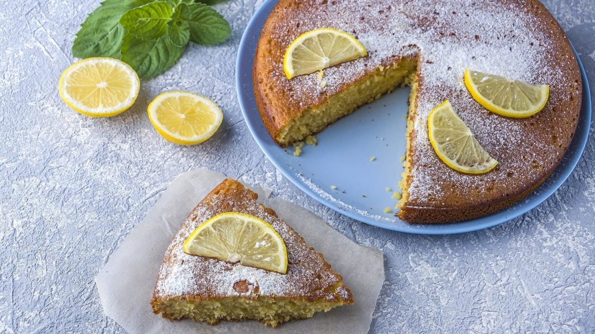 Vogelperspektive: Ein runder Grießkuchen mit Zitronen steht auf einer hellen Platte und diese auf einem hellen Untergrund. der Kuchen ist angeschnitten. Ein Stück liegt auf einem teller neben der Kuchenplatte.