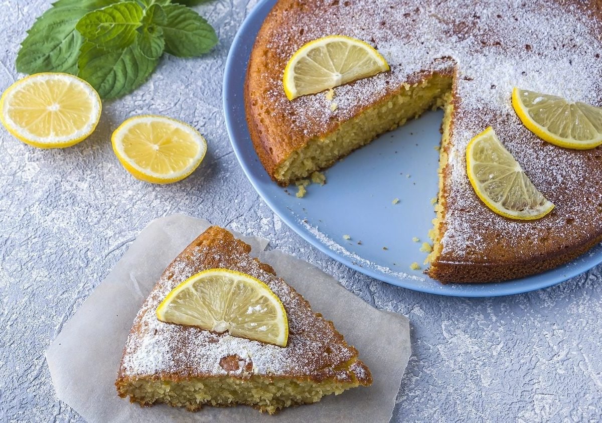 Vogelperspektive: Ein runder Grießkuchen mit Zitronen steht auf einer hellen Platte und diese auf einem hellen Untergrund. der Kuchen ist angeschnitten. Ein Stück liegt auf einem teller neben der Kuchenplatte.