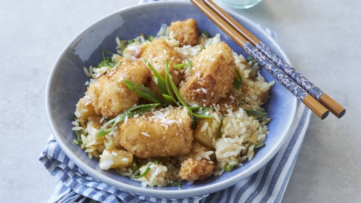 Draufsicht: Grauer Untergrund auf dem eine Schüssel auf einer blau-weißen Stoffservierte steht. In der Schüssel Kokos-Fisch-Nuggets mit Ananas-Reis.