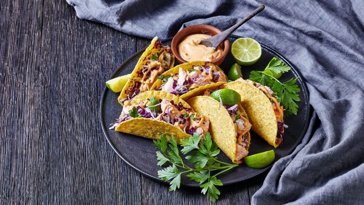 Draufsicht: Dunkler Untergrund mit einem grauen Geschirrtuch auf der rechten Seite. In der Bildmitte ein Teller mit fünf Lachs-Tacos mit Rotkohl und scharfer Joghurt-Sauce. Dazu frischer Koriander und Limette.