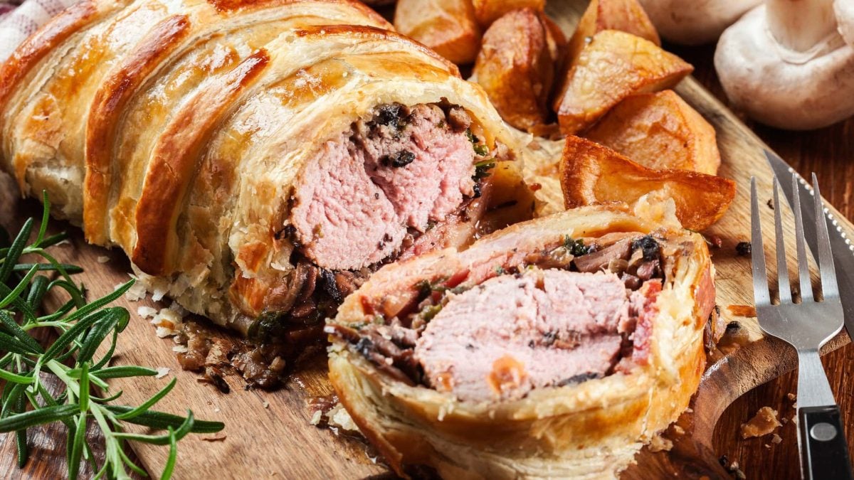 Schweinelende im Blätterteig mit Bacon und Pilzen aufgeschnitten auf Holzbrett. Daneben Rosmarinzweig und Kartoffelecken. Frontalansicht.