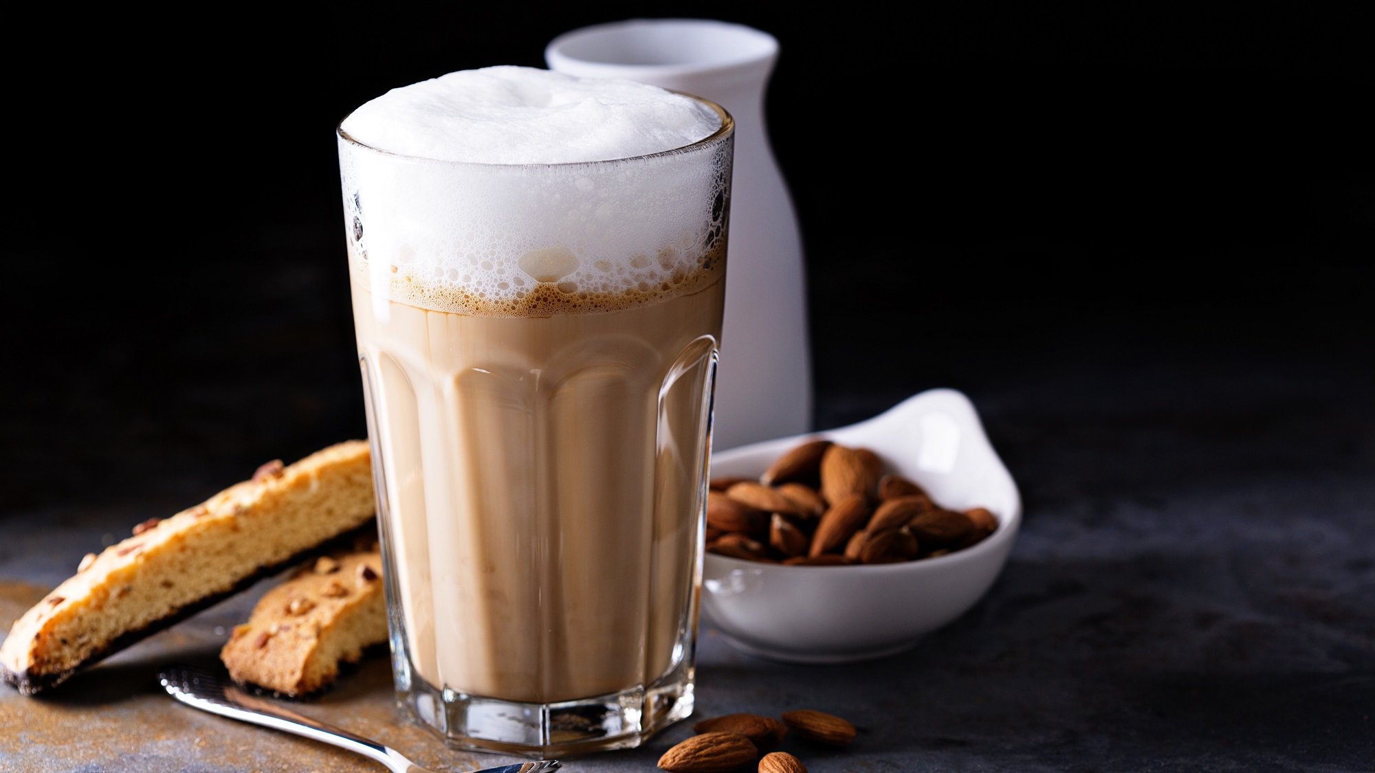 Ein Glas Mandel-Macchiato mit Milchschaum, daneben eine Schale Mandeln, ein Milchkännchen und Biscotti.