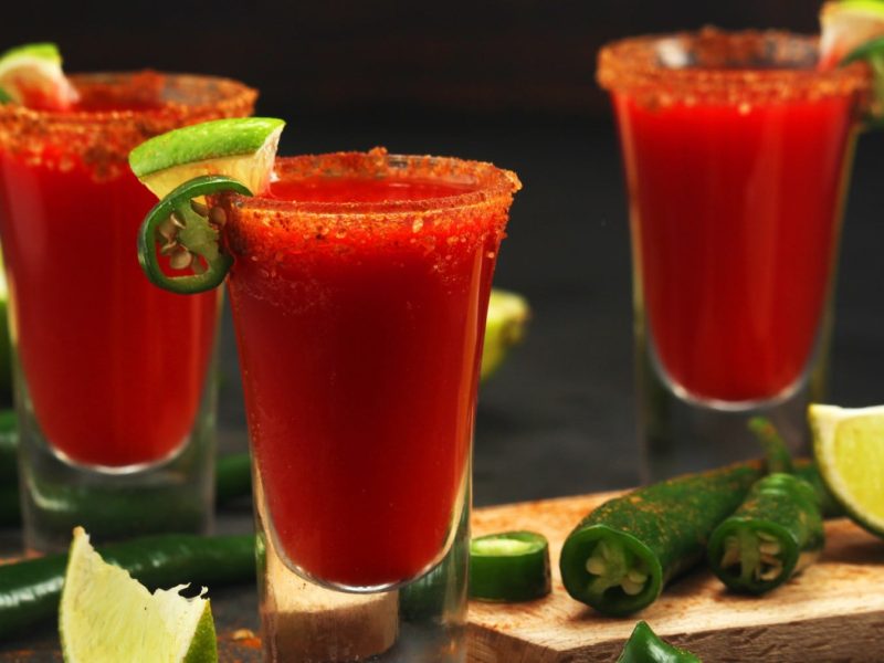 Drei Shotgläser mit Mexikaner Shot, drum herum Limetten und grüne Chilis vor dunklem Hintergrund.