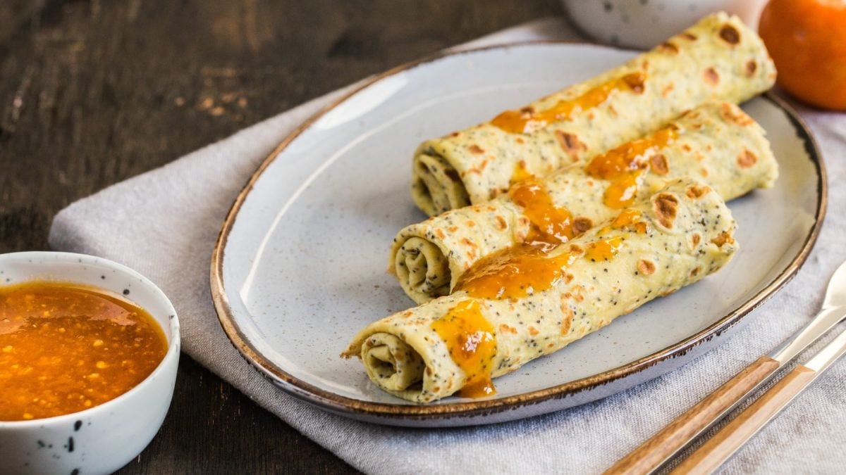 Ein weißer Teller mit den gerollten Mohn-Crepes auf dunklem Holztuntergrund. Daneben ein Schälchen mit Mandarinen-Marmelade.