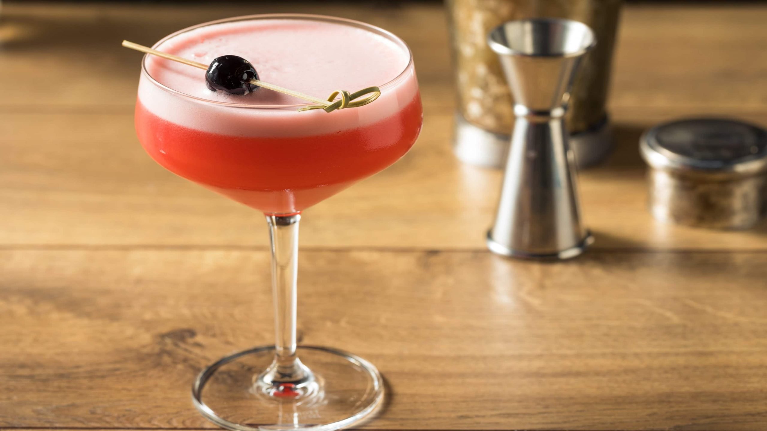 Negroni Sour in Cocktailglas auf Holz. Im Hintergrund Barmaß und Shaker. Frontalansicht.