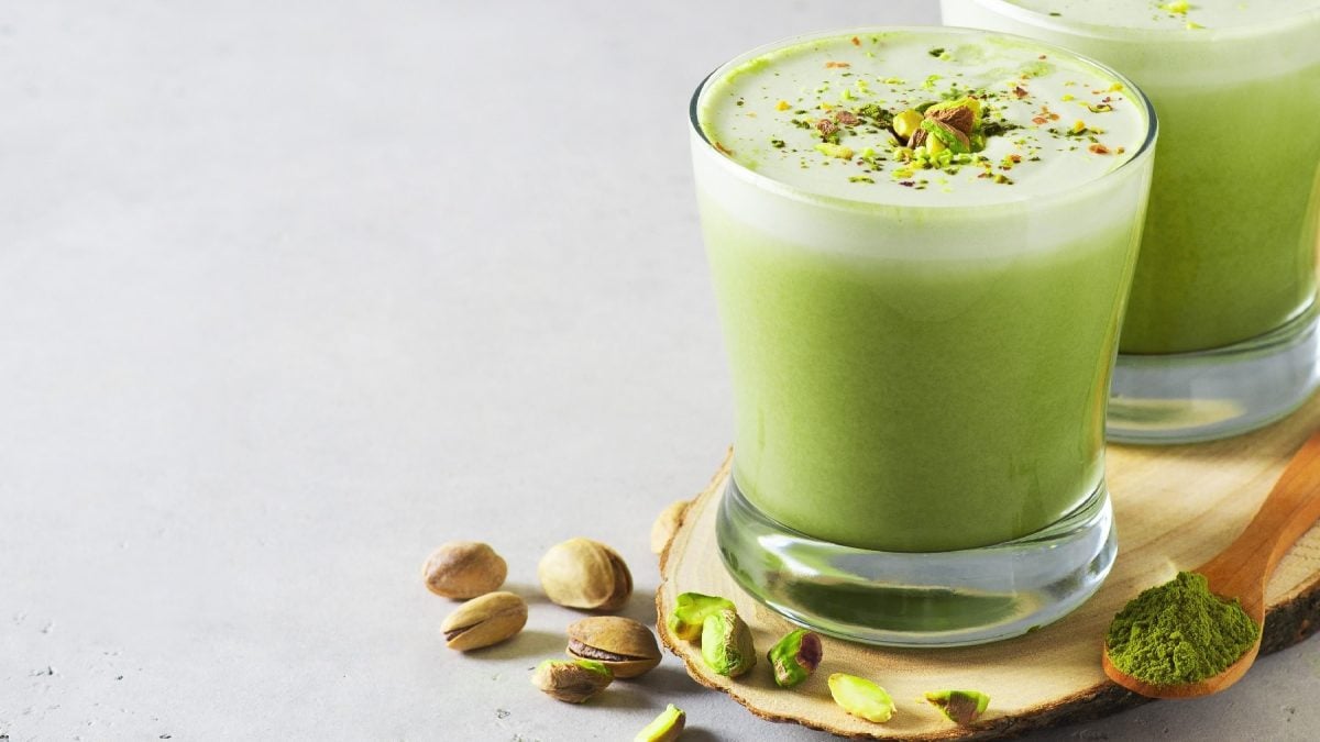 Zwei Gläser mit dem Pistazien-Matcha-Latte auf einem kleinen Holzbrett. Daneben ein Löffel mit dem Matchapulver und ein paar Pistazien.
