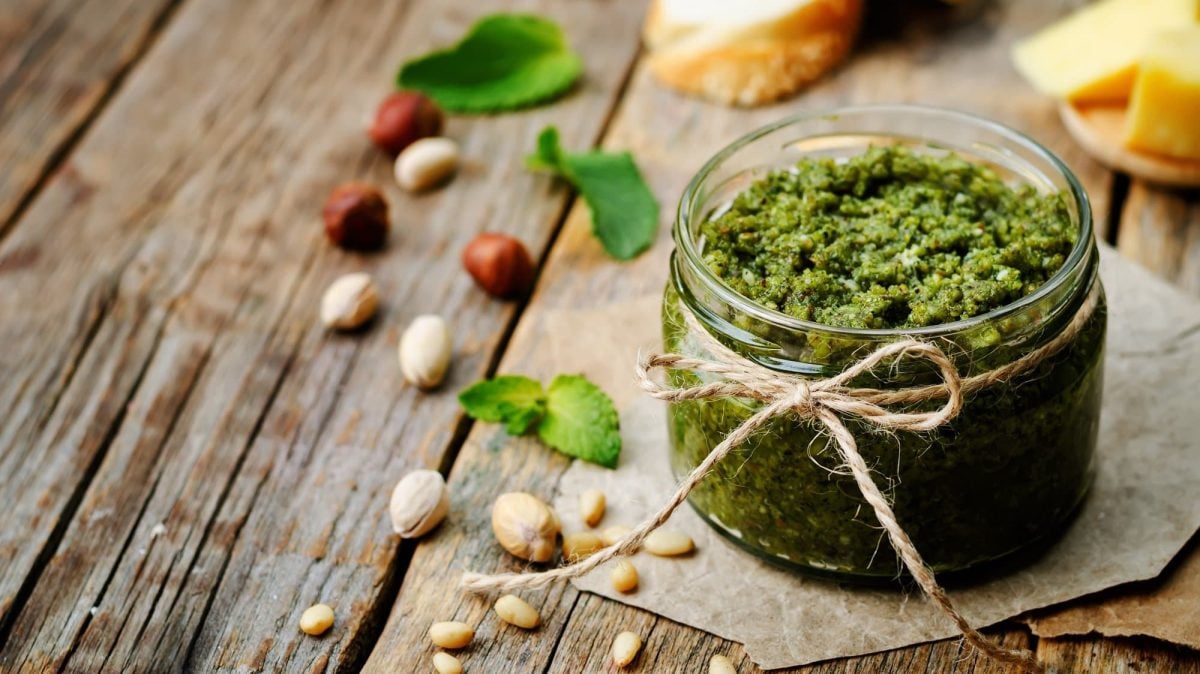 Ein Glas Pistazienpesto mit einer Schleife, daneben Pistazien und andere Nüsse.