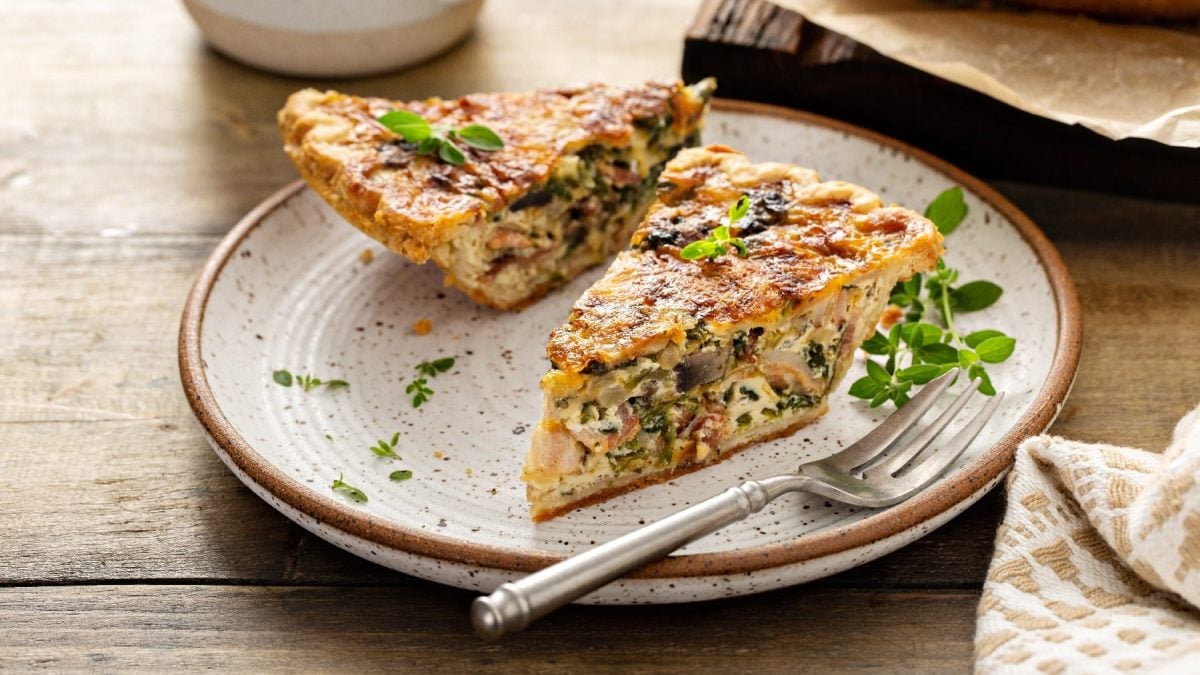 Ein weißer Teller mit braunem Rand und darauf ein Stück Quiche auf einem rustikalem Holztisch.